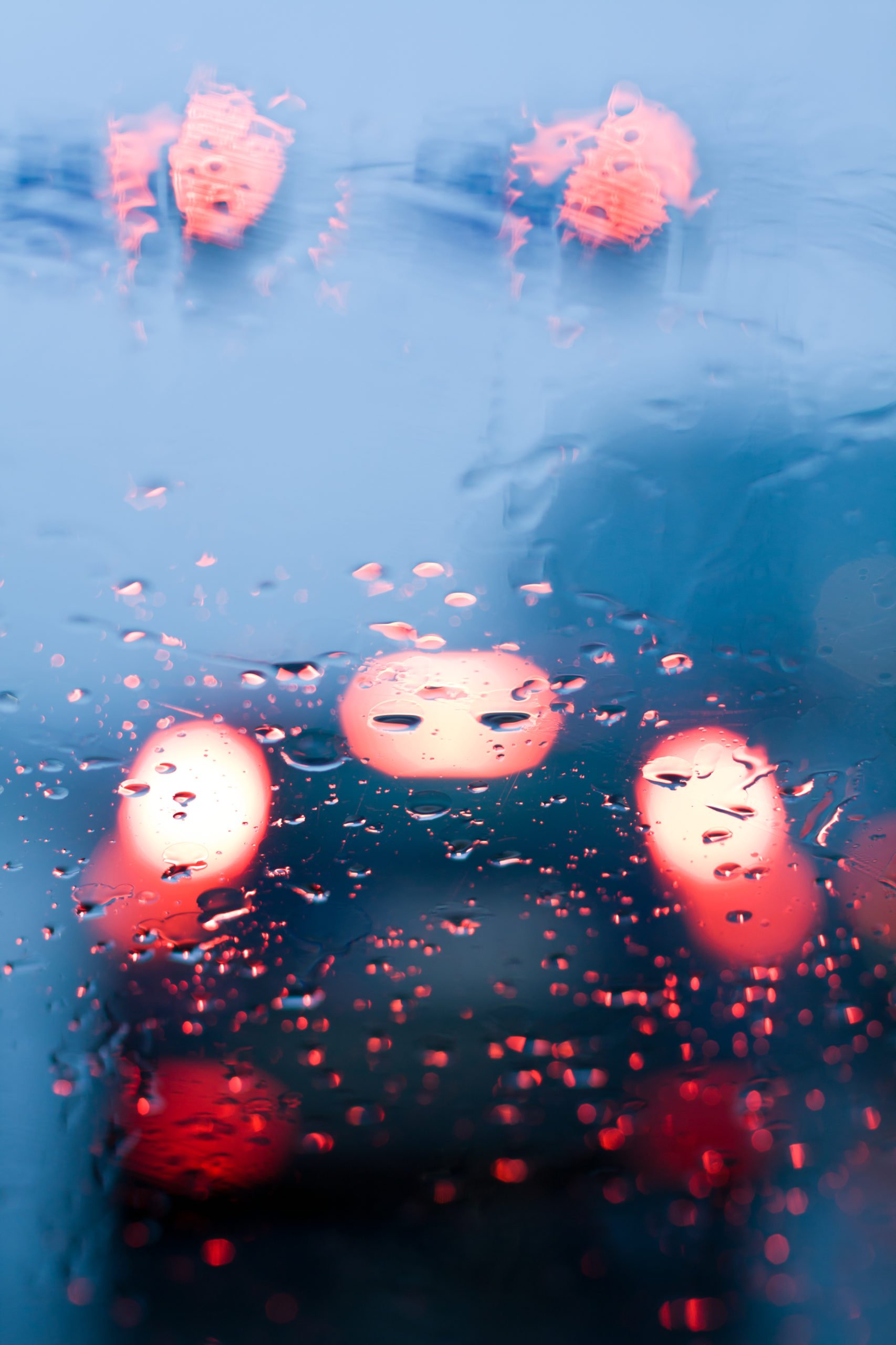 Car windshield with rain drops during storm and blurred stoplights.
Shallow depth of field with focus on center of the windshield with red lights.